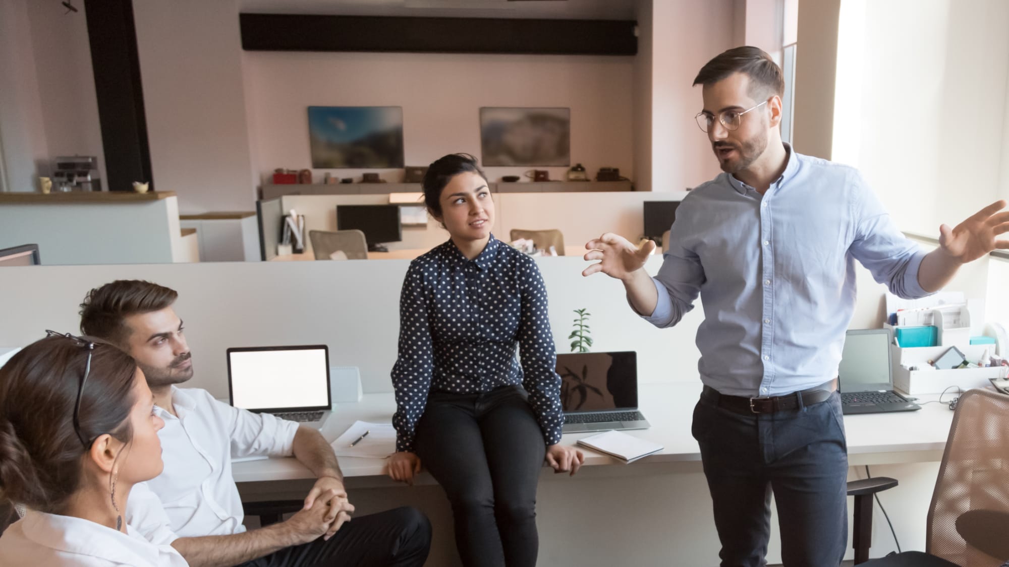 Four professionals meet in a bright office. A man in a blue shirt and glasses stands, gesturing widely while leading the discussion. Three colleagues sit nearby, listening intently to the speaker.